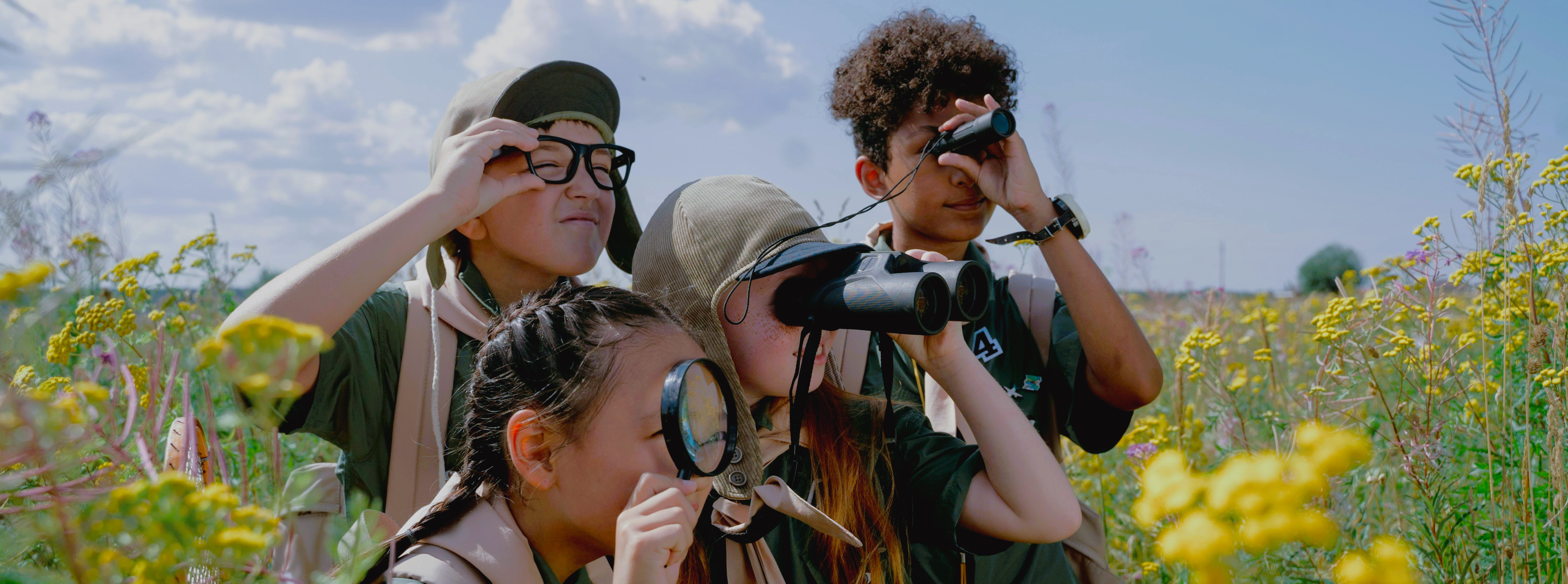 a group of scouts in a field using items such as binoculars and magnifying glasses to look at something off screen